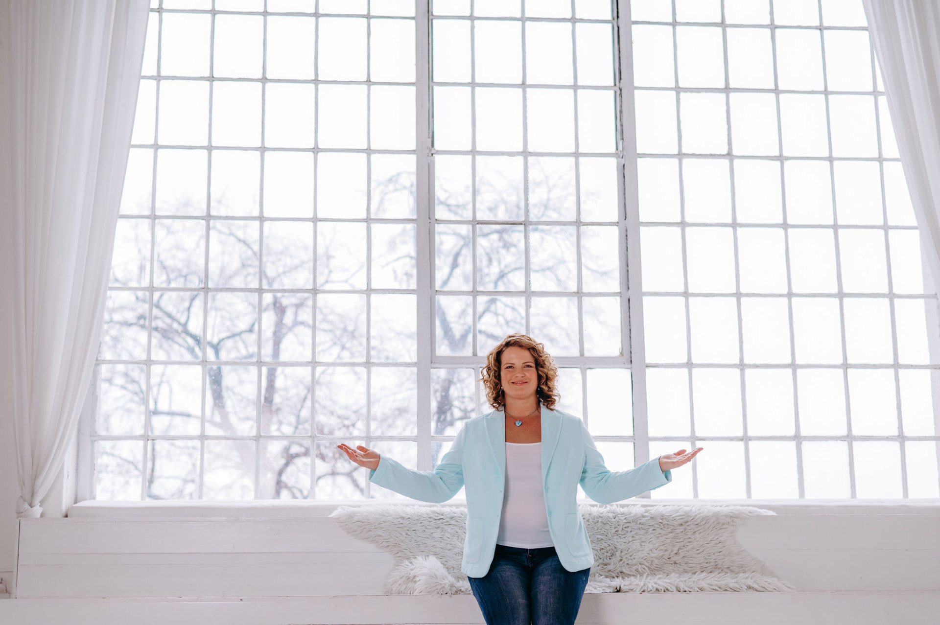 Petra Zlotá smiling with open arms in a light blue blazer before a tall bright window